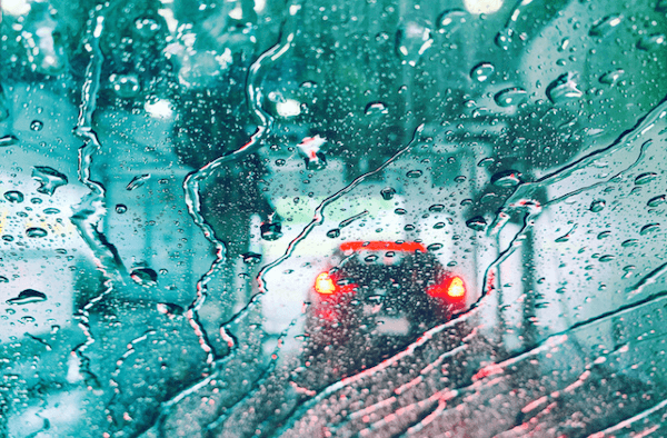 A red car is driving down a wet road in the rain.