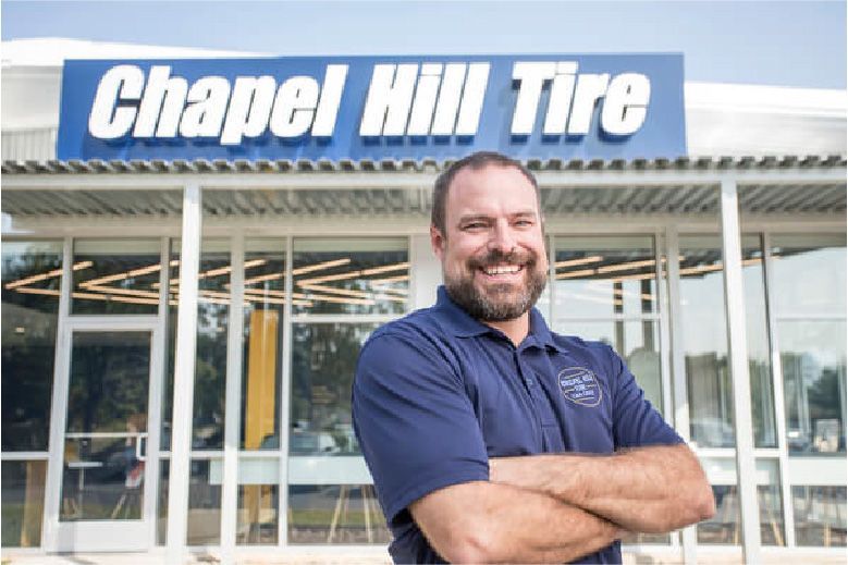 Man with beard smiles in front of Chapel Hill Tire storefront with blue sign.
