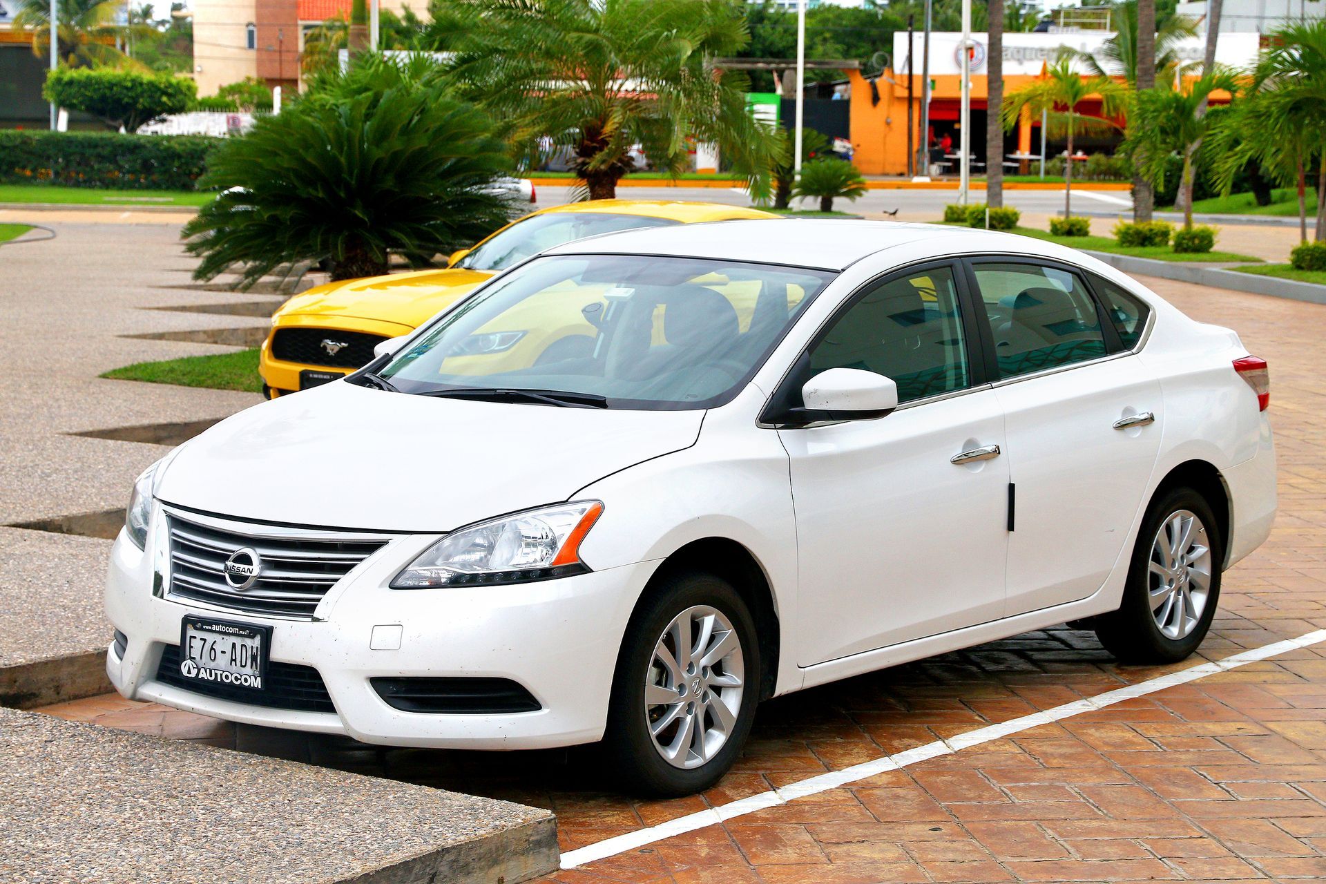 White Nissan Sentra parked in a paved lot, with a yellow car in the background; palms trees and a building are visible.
