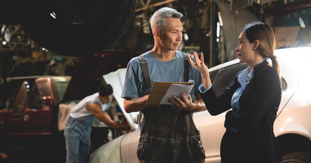 A man is talking to a woman in a garage while a man works on a car.