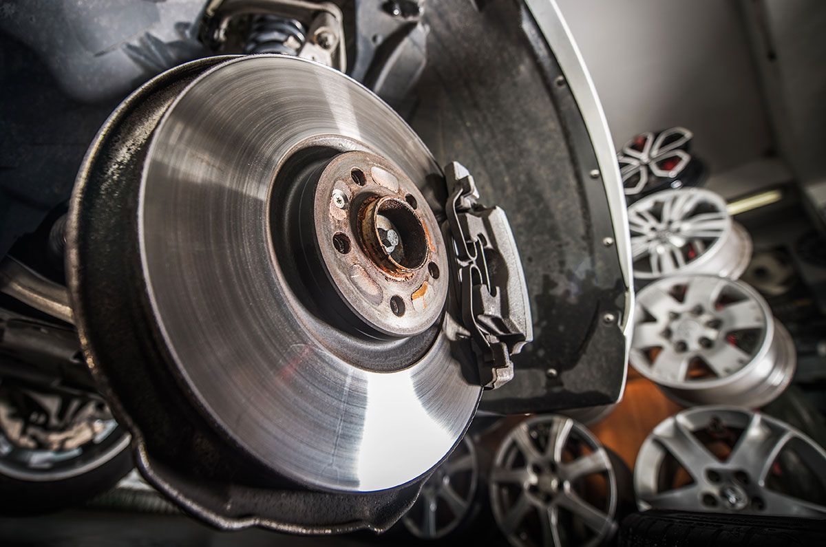 A car brake disc and caliper assembly in a garage, with several spare alloy wheels stacked in the blurred background.