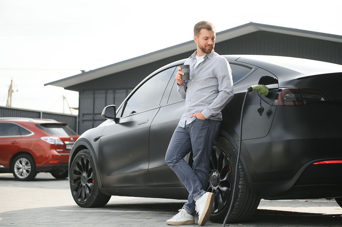 Man in casual wear leans against a black Tesla, holding a coffee cup, outdoors. Another car is parked behind.