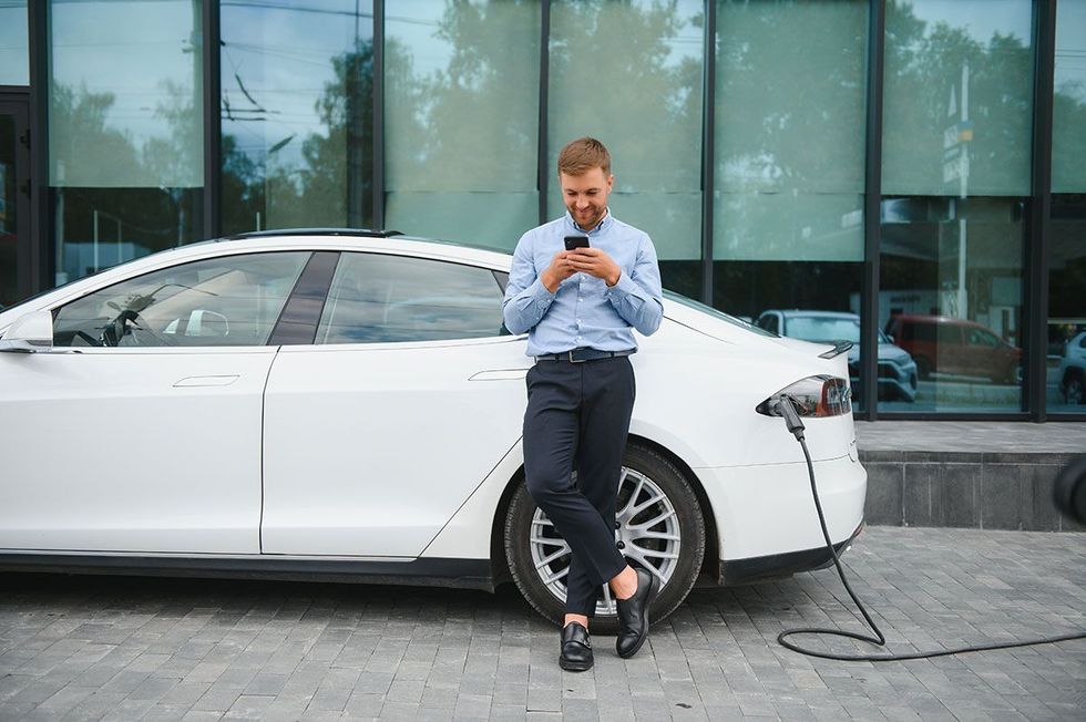 Man leaning against a white electric car while charging his phone outside a glass-front building