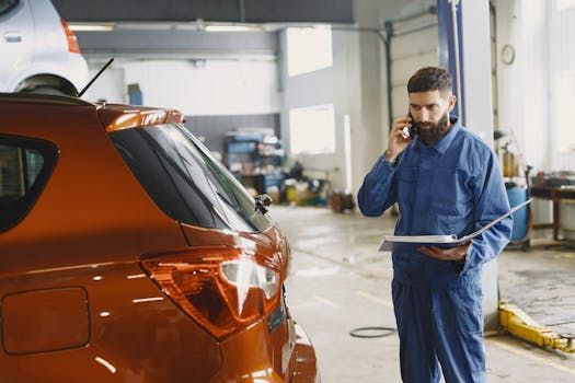 Mechanic in blue jumpsuit, talking on phone, holding paperwork near an orange car in a garage.
