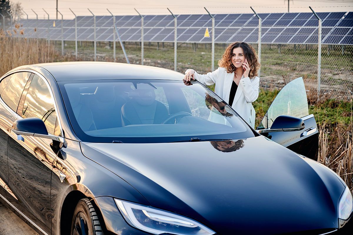Woman in white blazer leans on a black electric car, smiling while on a call, solar panels in the background.