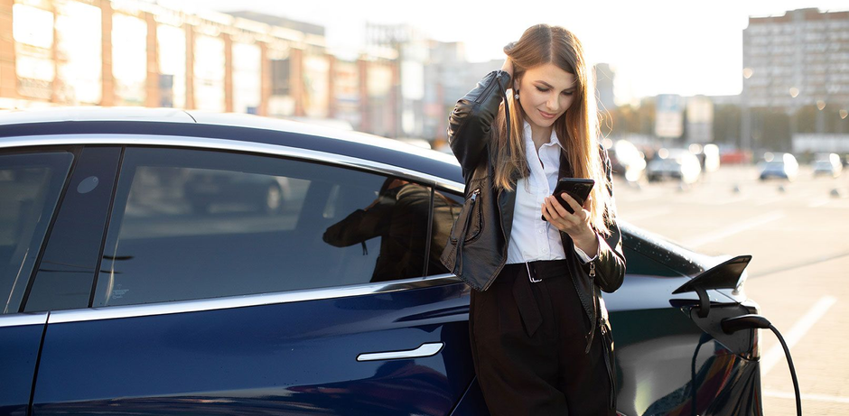 Man leaning against a white electric car while charging his phone outside a glass-front building