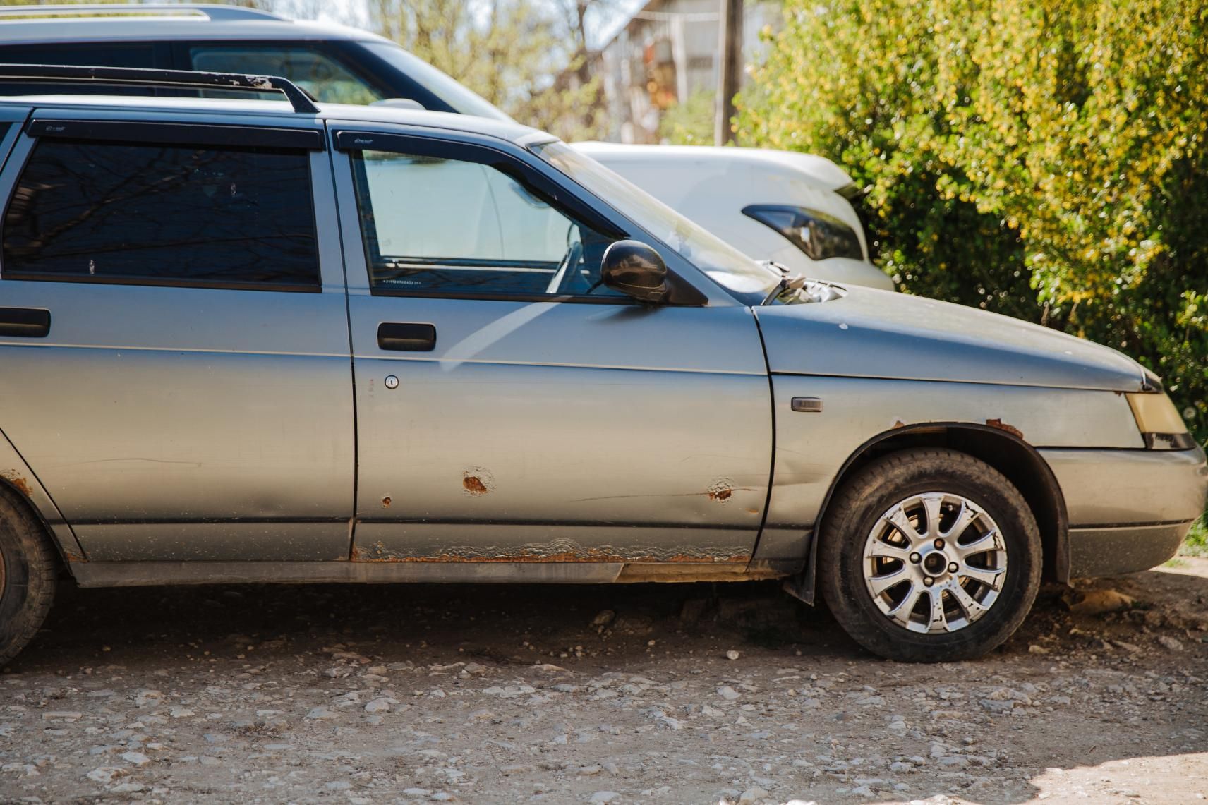 A silver car is parked in a gravel lot next to another car.