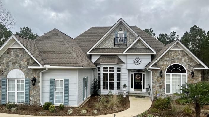 Large, two-story house with stone accents, light blue siding, and a curved walkway under a cloudy sky.
