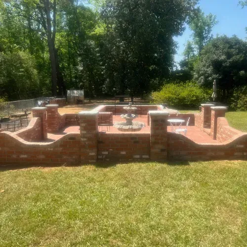 Brick patio with fountain and low walls, in a grassy yard, surrounded by trees.
