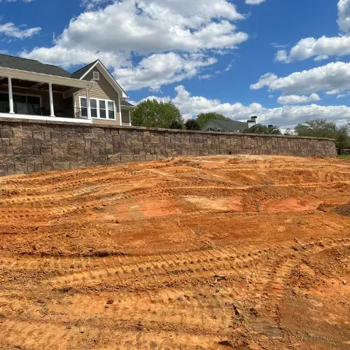 Brown retaining wall in front of a house, with exposed reddish earth and a blue sky with clouds.