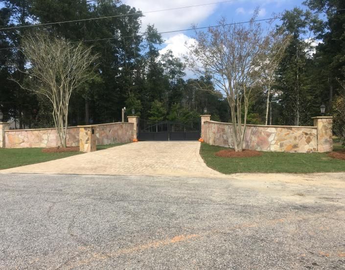 Stone wall entrance with a gate and gravel driveway. Trees border the entrance; a green lawn.