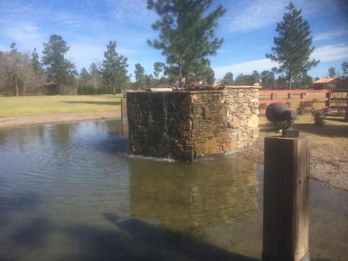 Water fountain with stone wall, flowing into a pond outdoors. Trees and blue sky visible.