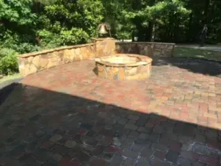 Brick patio with stone fire pit and retaining wall, surrounded by trees.