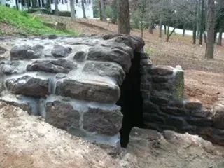 Stone tunnel entrance in a hillside, surrounded by dirt and grass, trees in the background.