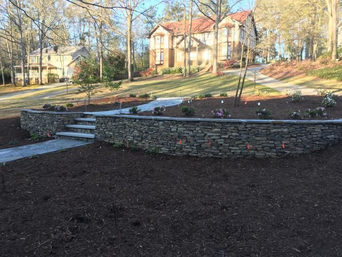Stone retaining wall with steps, leading to a landscaped area and house on a hill. Brown mulch covers the ground.