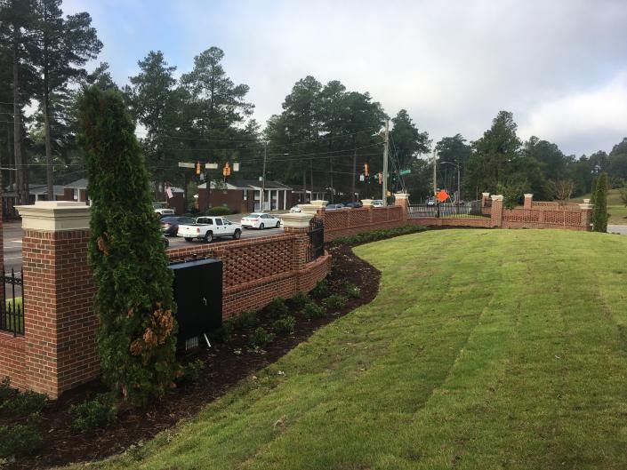 Brick entrance with a curved wall, green lawn, and road with cars.