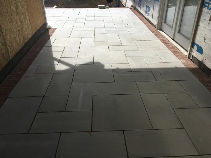Gray stone patio with brick border next to a wall and doorway; sunlight casts shadows.