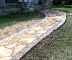 Stone walkway leading to a house, bordered by brick and surrounded by grass.
