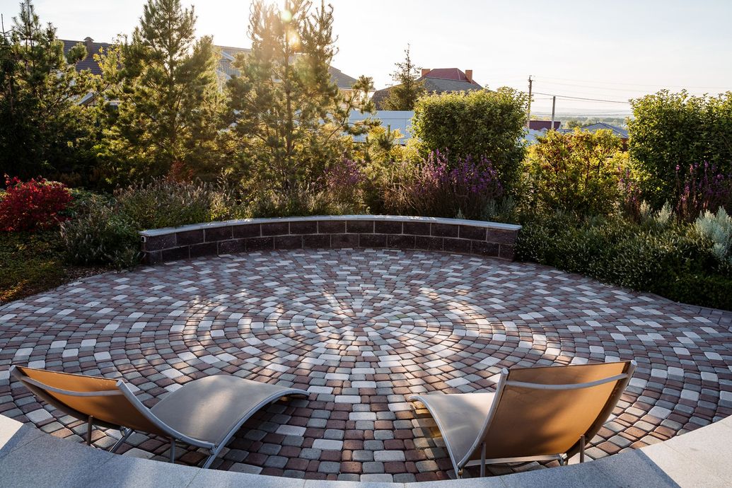 Two lounge chairs on a brick patio overlook a garden with colorful plants; sunlit trees in the background.
