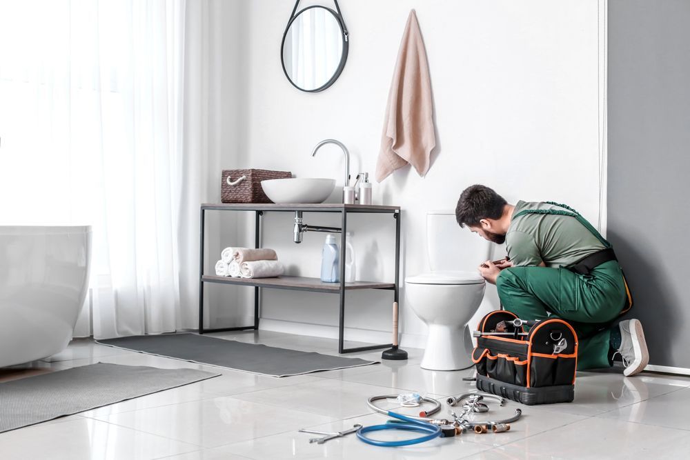 A plumber in green coveralls kneels on a bathroom floor, repairing a white toilet with tools nearby.