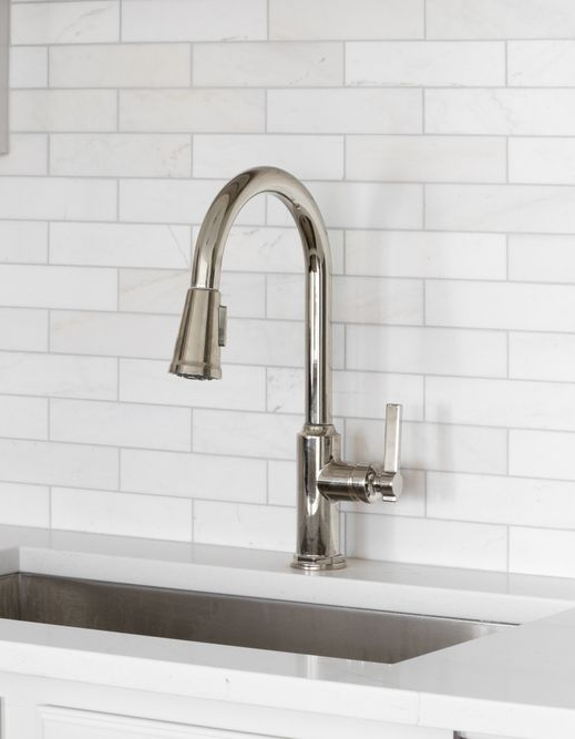 A modern, stainless steel high-arc pull-down kitchen faucet installed on a white countertop with a white tile backsplash.