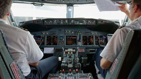 Två piloter i cockpit, en läser papper, instrument och reglage synliga.
