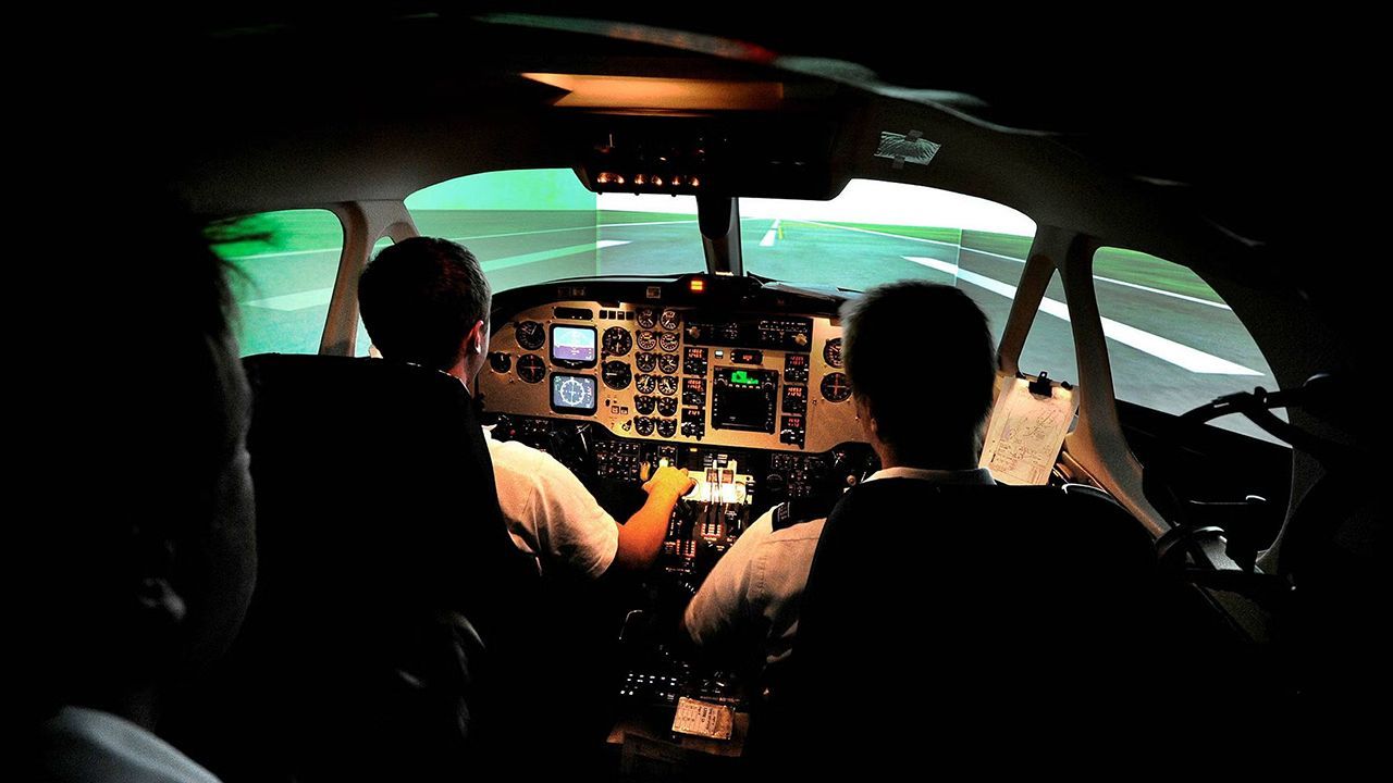 Inuti en cockpit: Två personer som styr ett flygplan och tittar på de upplysta reglagen och banan framför.