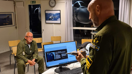 Två män i gröna overaller i ett rum, en presenterar sig med ett modellflygplan nära en datorskärm som visar foton.