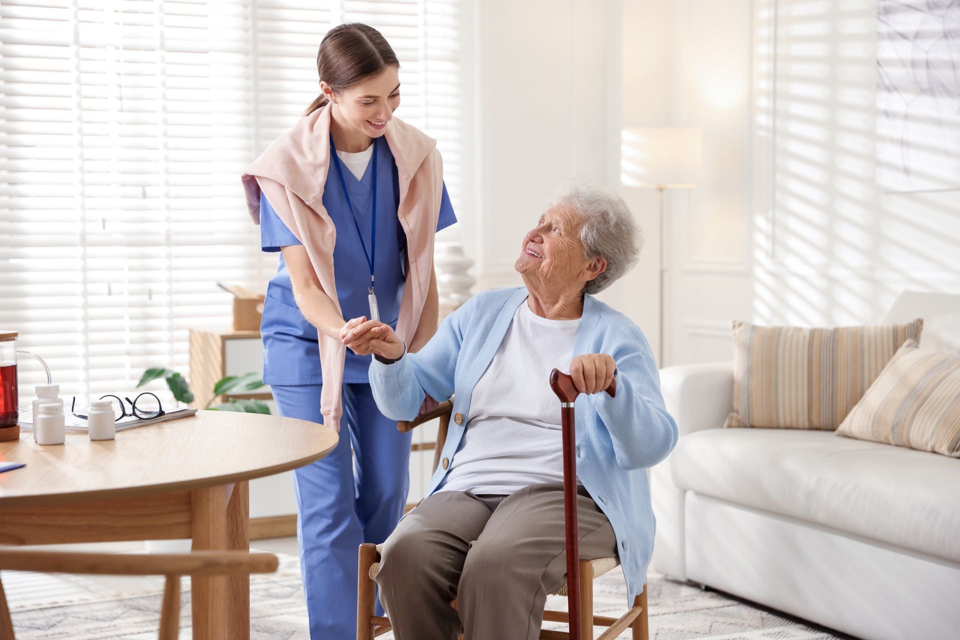 A caregiver in blue scrubs holding the hand of an older person seated with a cane in a bright, modern living room. A caregiver in blue scrubs holding the hand of an older person seated with a cane in a bright, modern living room.