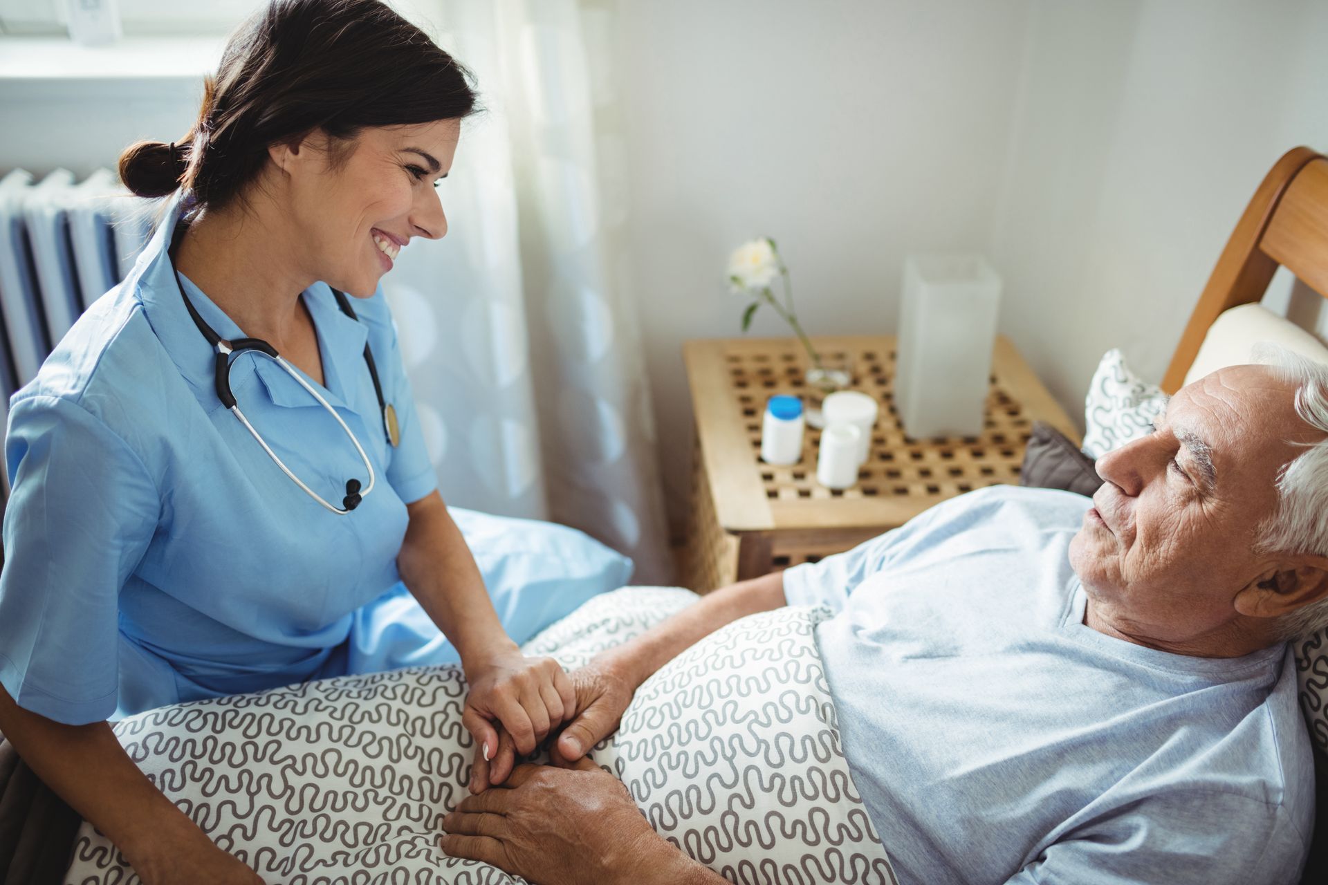 A healthcare worker in blue scrubs holds the hands of a patient lying in bed, smiling during a supportive interaction.