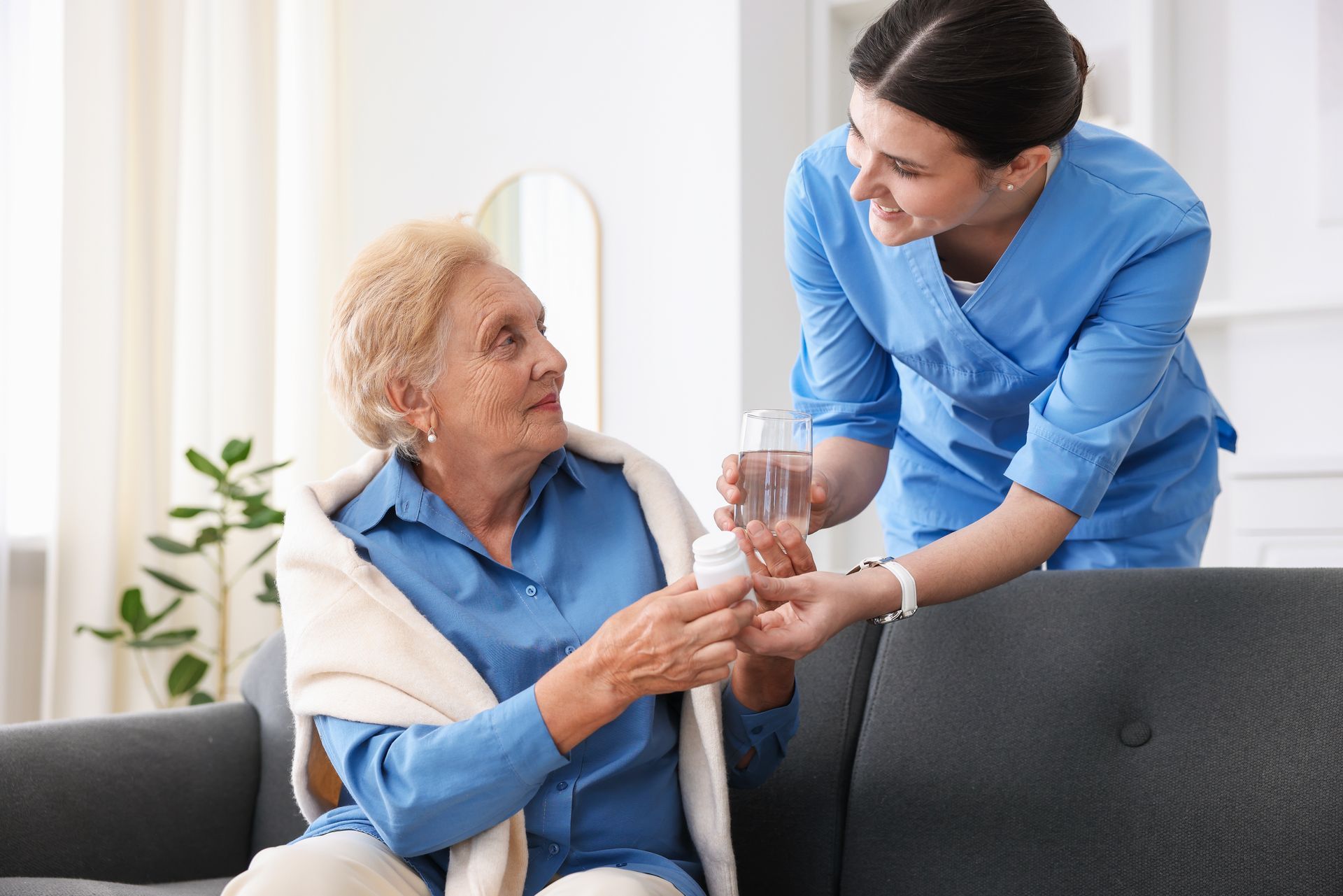 A person in a blue uniform hands a small pill container and a glass of water to an individual sitting on a gray sofa. A person in a blue uniform hands a small pill container and a glass of water to an individual sitting on a gray sofa.