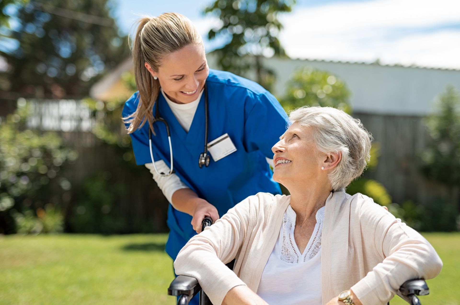 A healthcare worker in blue scrubs smiling at a person in a wheelchair on a sunny outdoor lawn.