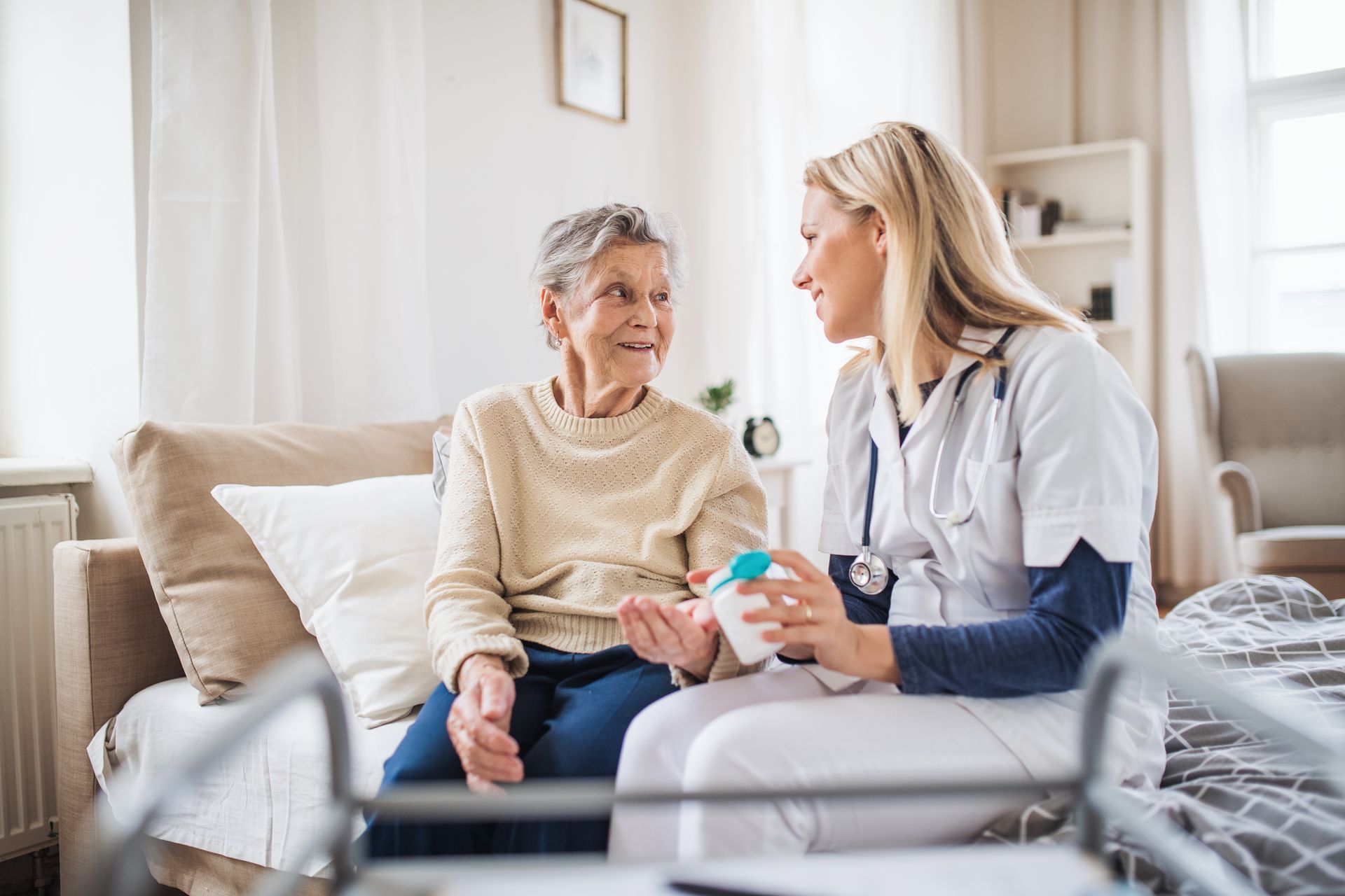A healthcare worker in uniform sits on a bed and helps a patient take medication from a bottle.