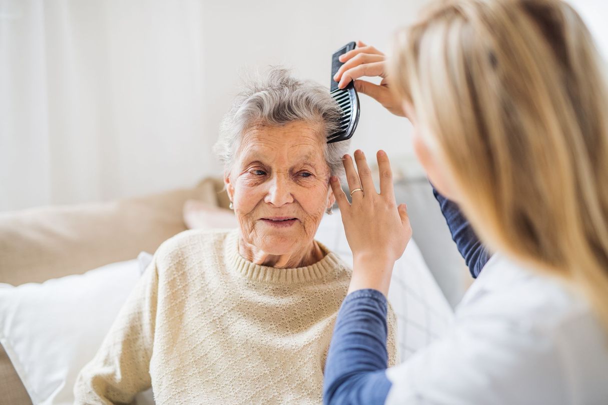 A caregiver combs the hair of a person sitting on a bed in a bright, indoor setting. A caregiver combs the hair of a person sitting on a bed in a bright, indoor setting.