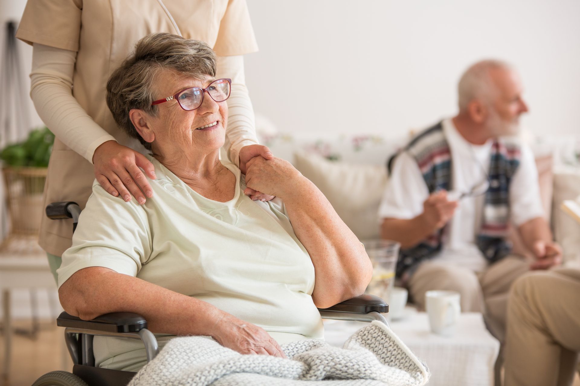 A caregiver stands behind a smiling person in a wheelchair, with another person sitting on a sofa in the background.