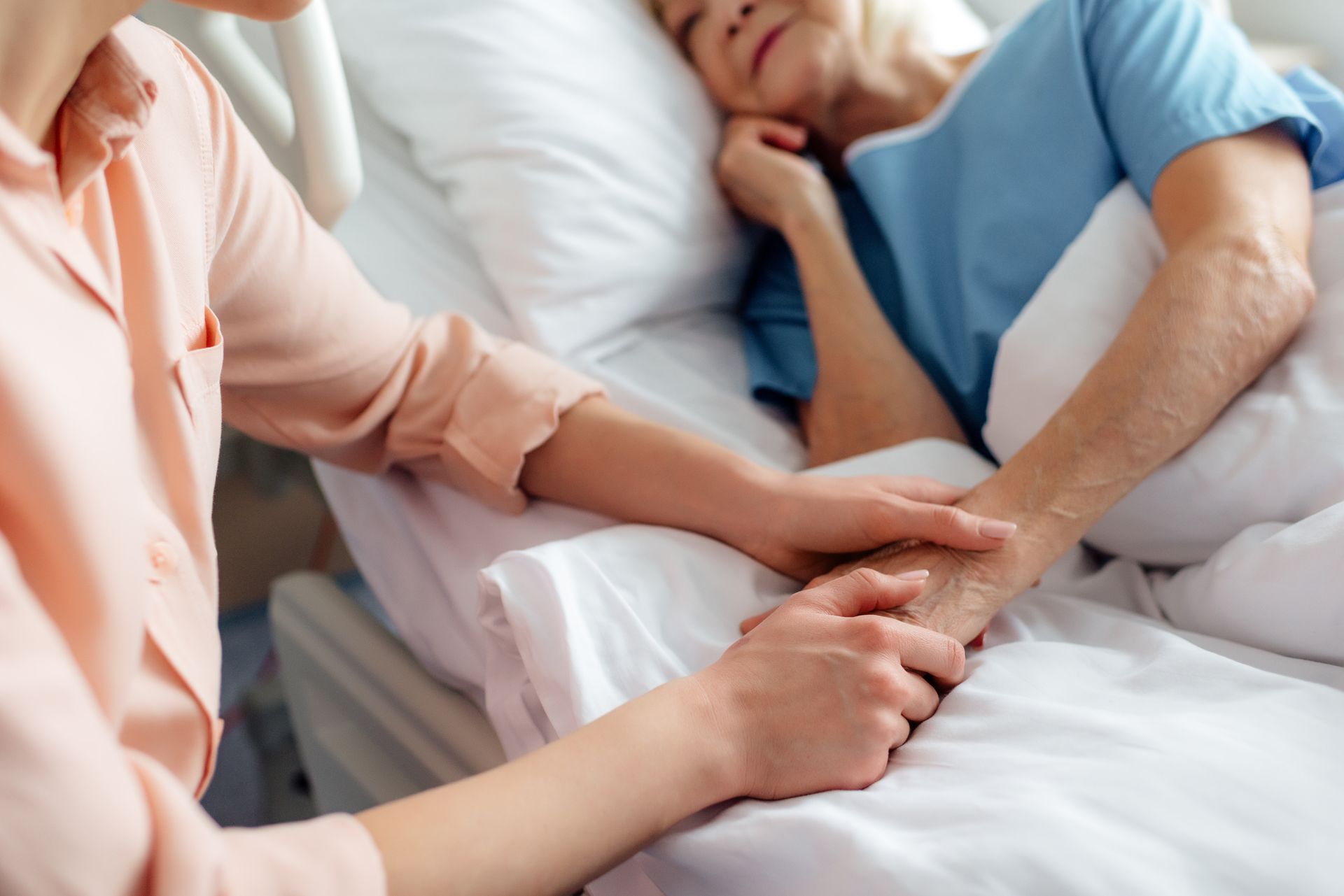 A person in a peach shirt holds the hand of someone lying in a hospital bed, offering comfort and support.