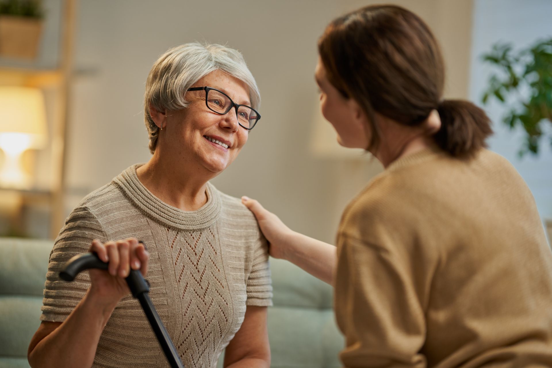 A person with a cane smiles as someone places a supportive hand on their shoulder in a warm, indoor setting.