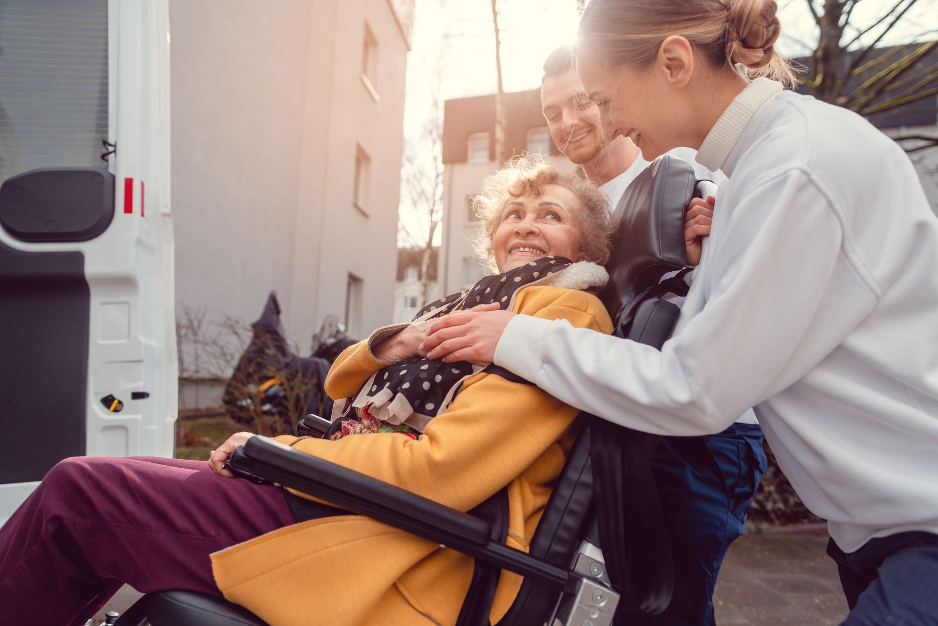 A caregiver in a white top smiles while helping an individual in a yellow jacket and wheelchair outdoors near a van.