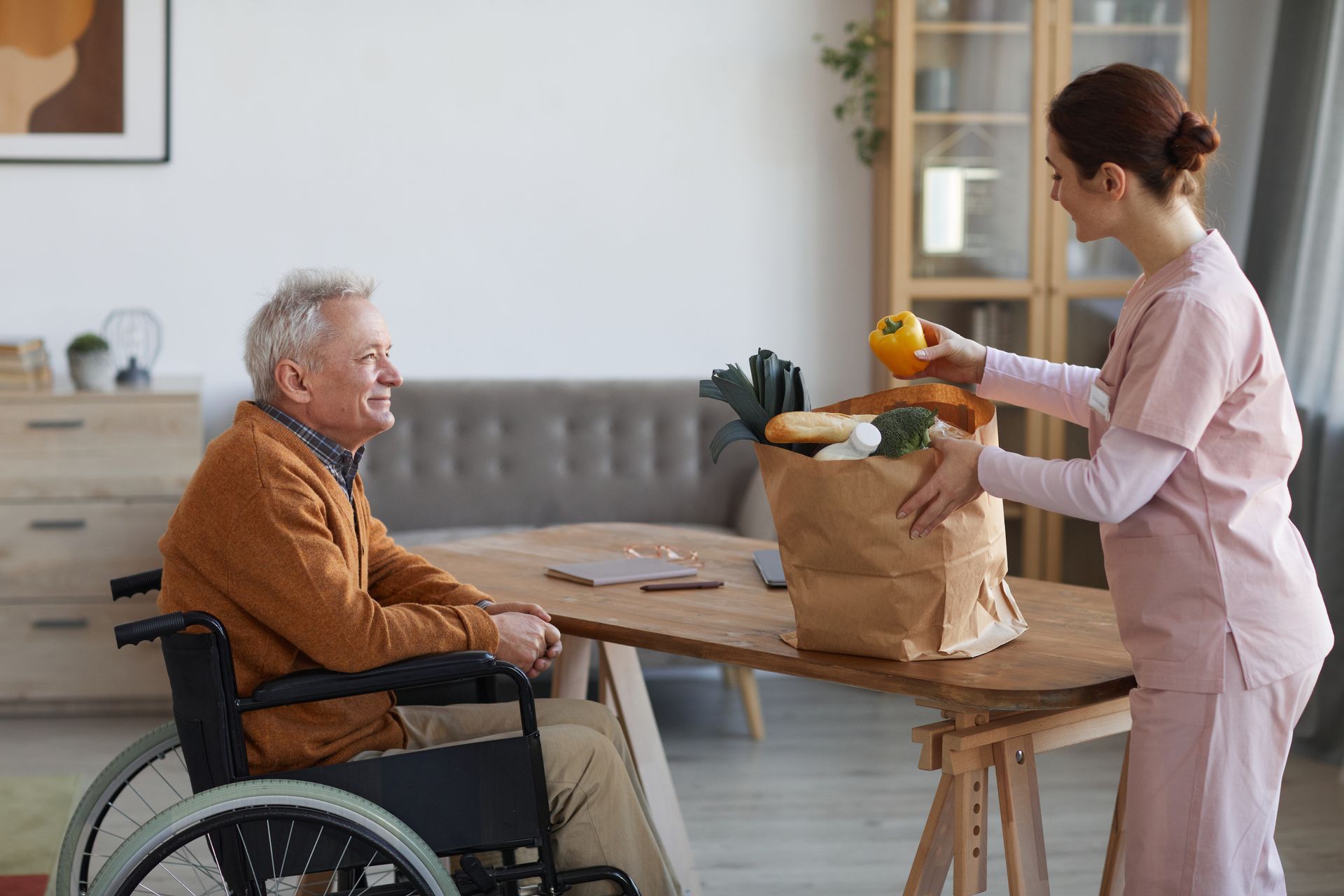 A caregiver in scrubs stands by a table, showing a yellow bell pepper from a grocery bag to a person seated in a wheelchair.