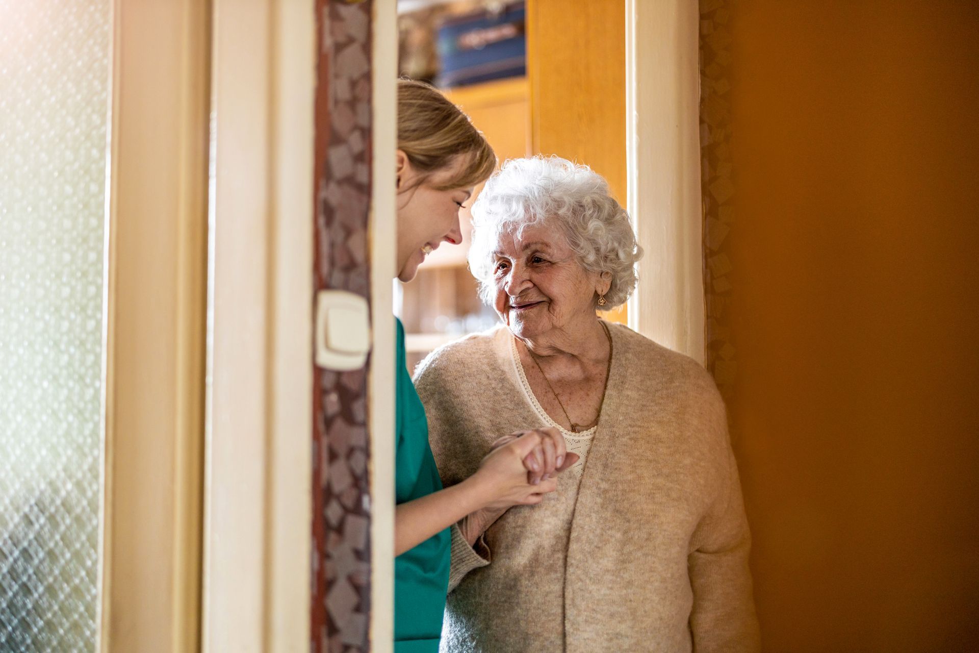 A care provider wearing teal scrubs gently holds hands with a smiling individual in a beige cardigan inside a home.