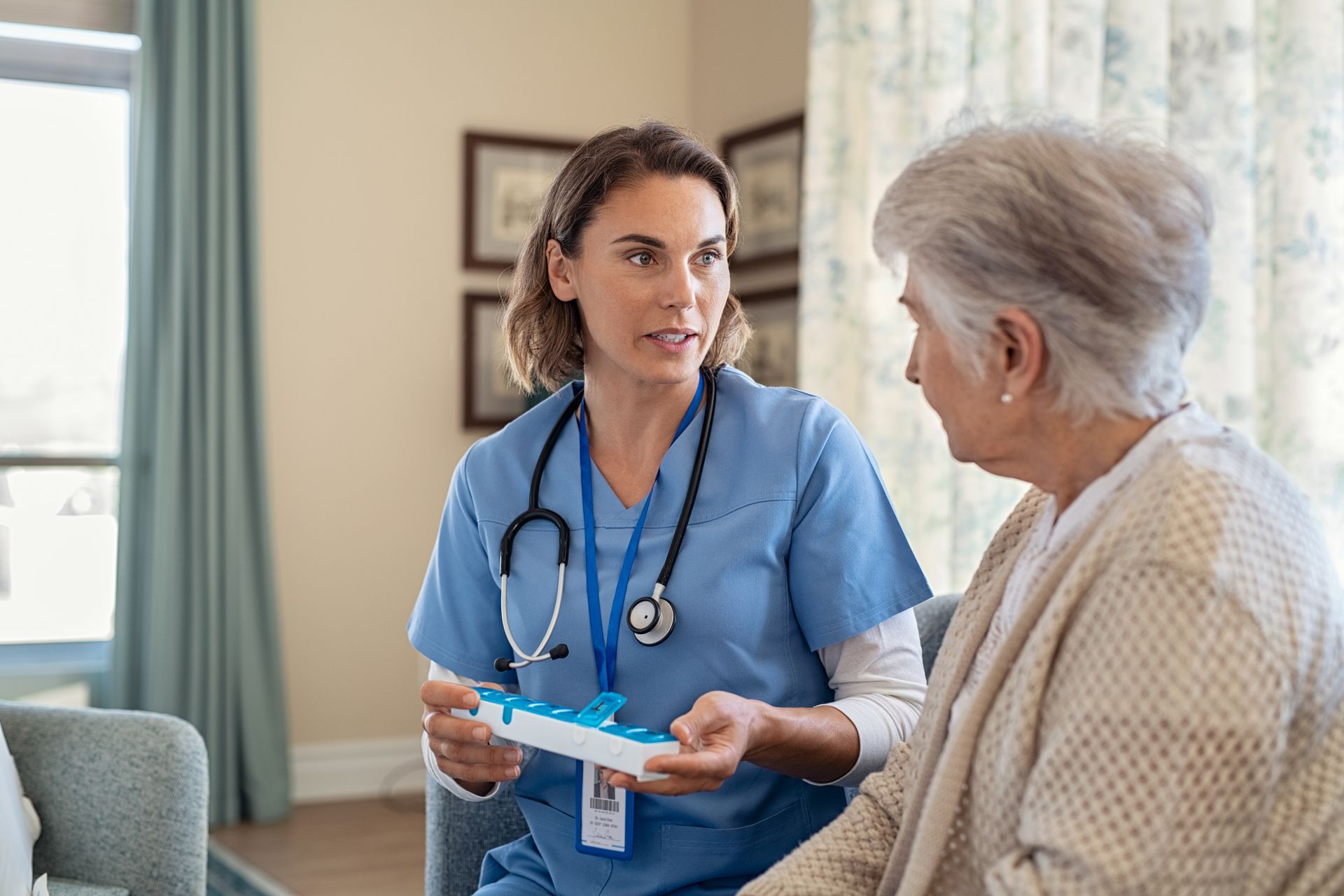A healthcare worker in blue scrubs explains a weekly pill organizer to a patient in a home setting.