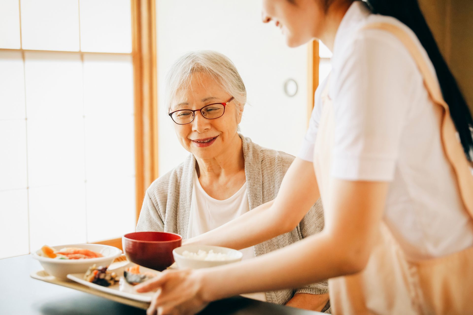 A caregiver serves a tray with a bowl of rice, soup, and side dishes to a person seated at a table.