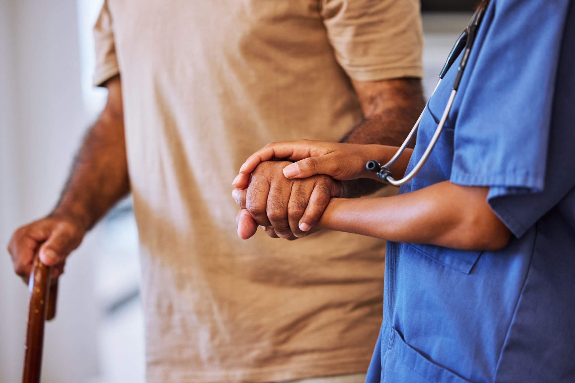 A caregiver in blue scrubs holding the hand of an individual leaning on a wooden cane.