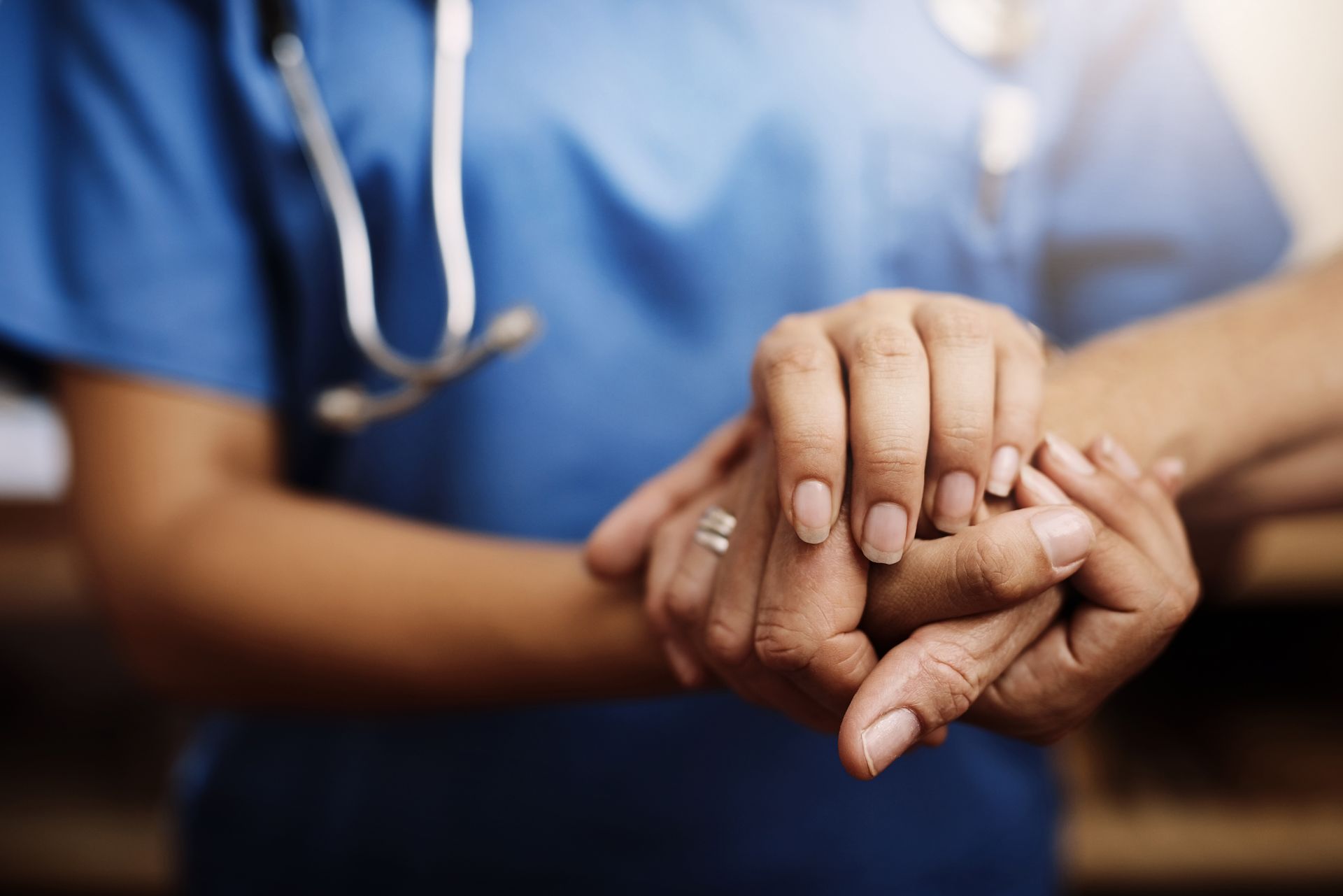 A healthcare worker in blue scrubs holding a person's hands in a gesture of comfort and support.