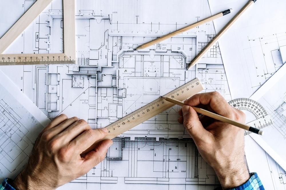 Hands measuring a blueprint with a ruler and pencil, surrounded by drafting tools and floor plans.