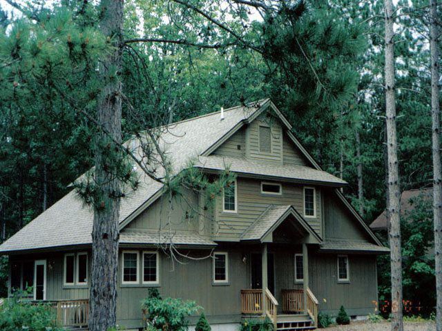Two-story, green house nestled amongst tall trees; features a covered porch and numerous windows.