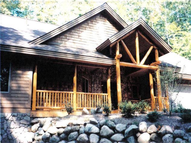 Log cabin-style house with wooden porch, stone foundation, and wood shingle roof.
