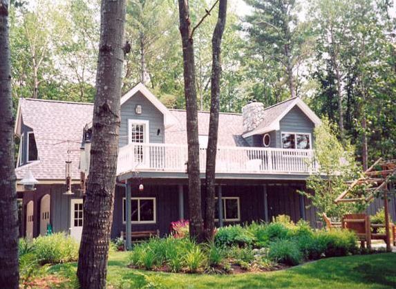 Gray house with balcony, surrounded by trees and a garden in front.