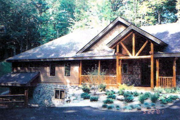 Rustic brown house with stone foundation, porch, and timber accents; surrounded by trees and shrubs.