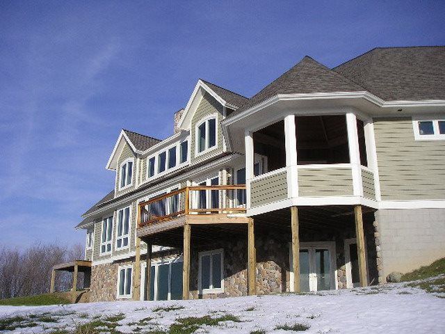 Multi-story house with decks, light green siding, and stone base, on a snowy hill, under blue sky.